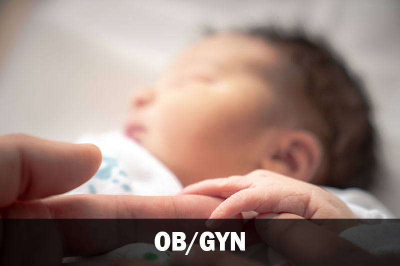A newborn baby touching holding an adults finger. There is a text overlay that says "OB/GYN."