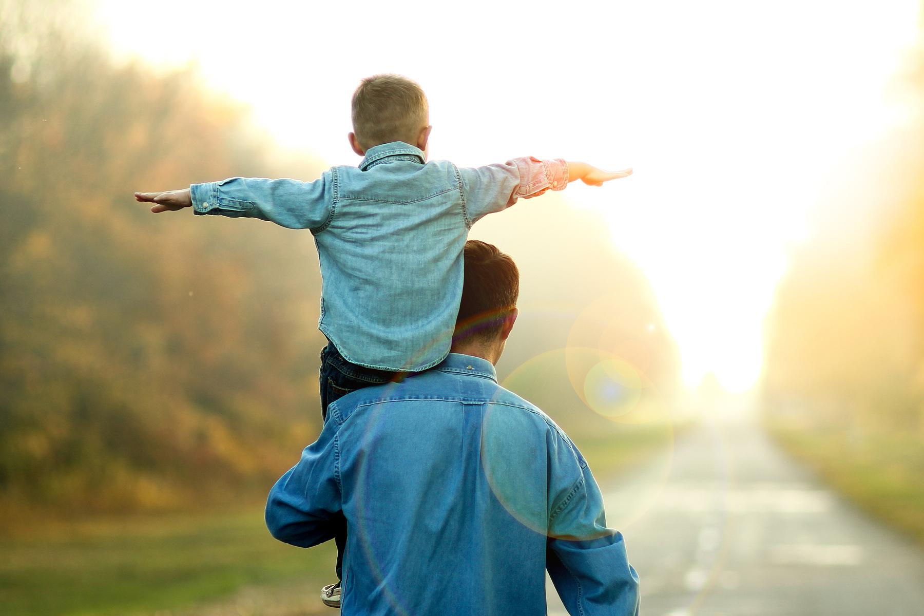 A boy riding on a man's shoulder with his arms spread out like wings.