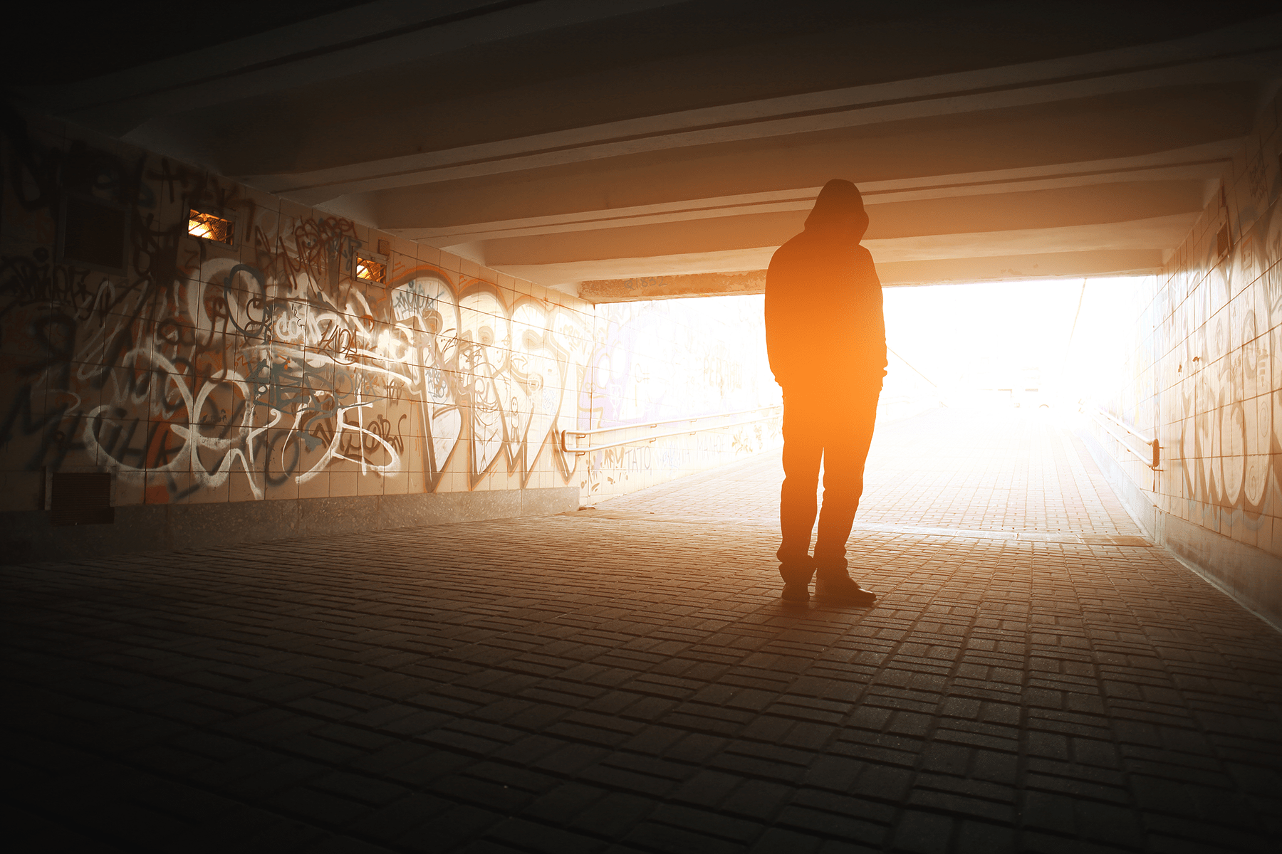 A hooded figure stands under a bridge with graffiti on the wall and the sun shining down into the structure.
