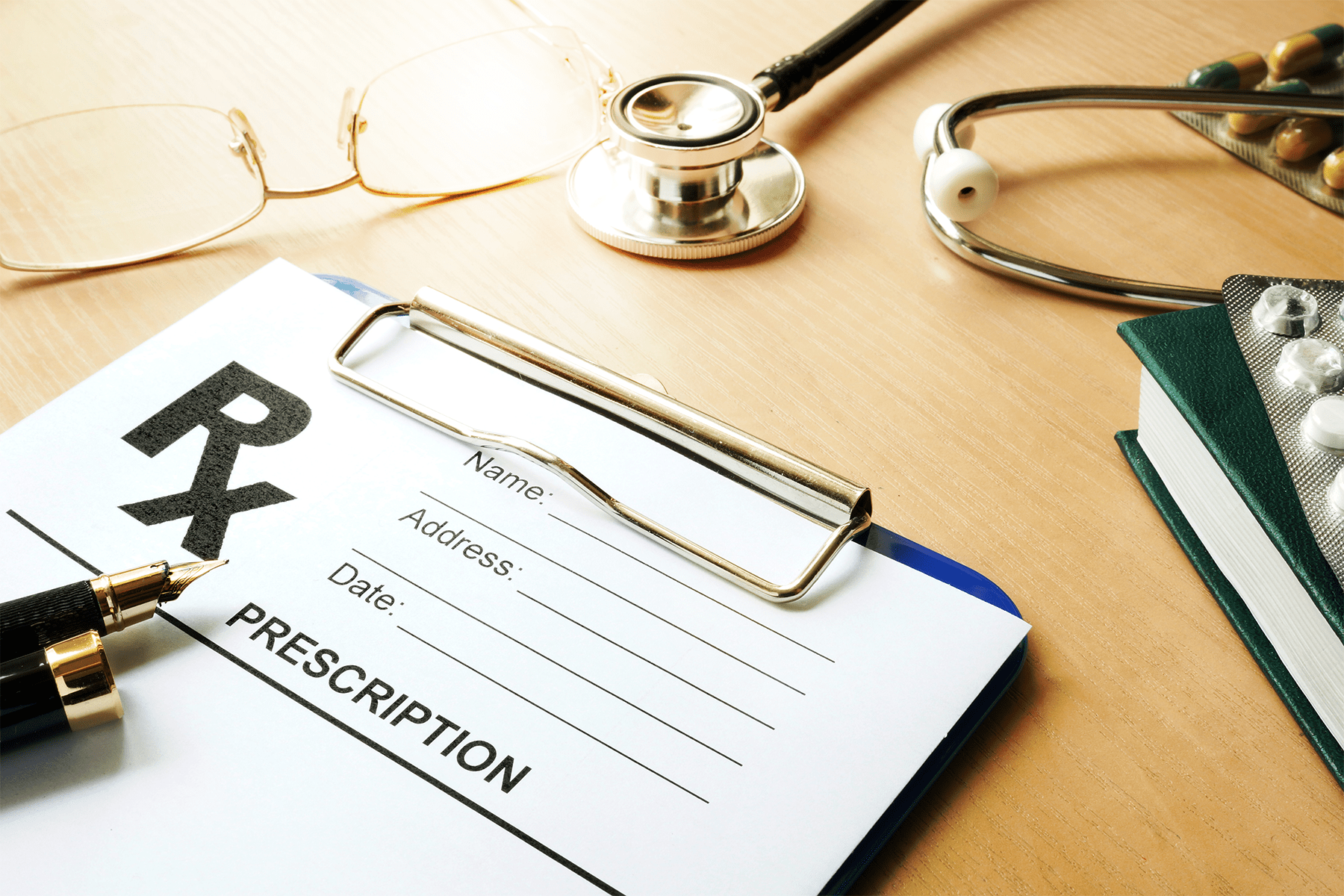 A prescription pad, stethoscope, glasses, and some pills lie on a table.