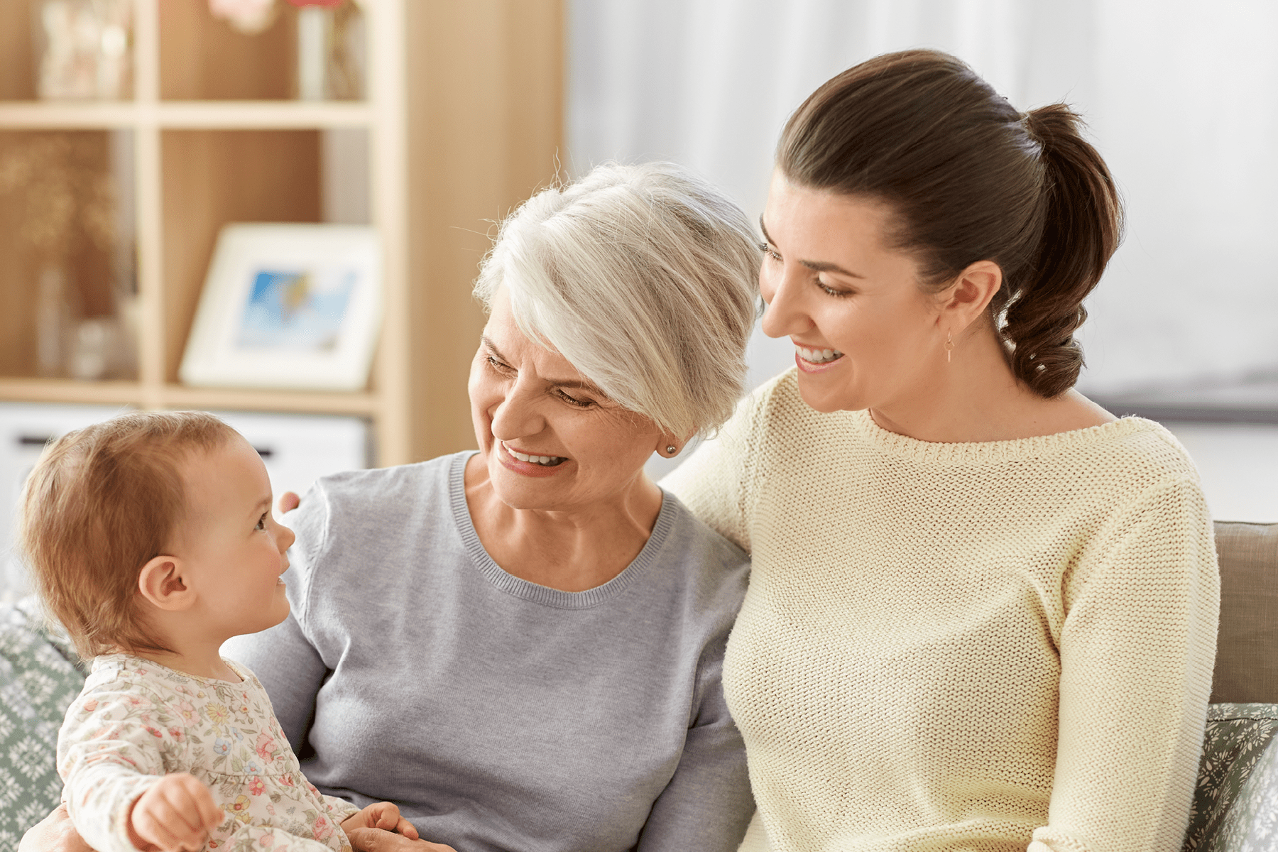 Two woman sit on the couch they are smiling at a baby that one of the women is holding.