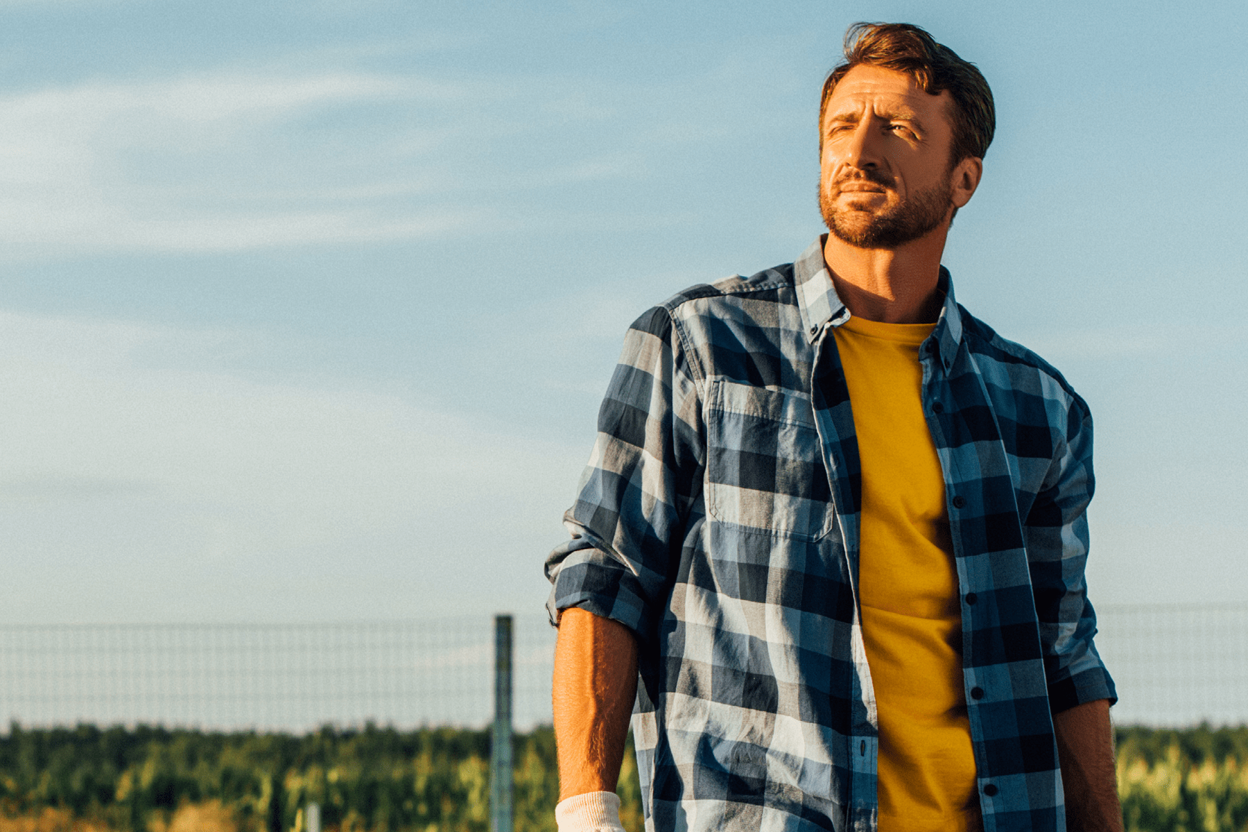 A man wearing a flannel shirt stands in the foreground with a fence and wide open sky in the background.