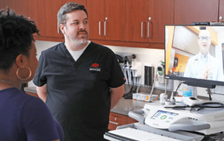 A doctor appears virtually to speak with a nurse and a patient in an examination room.