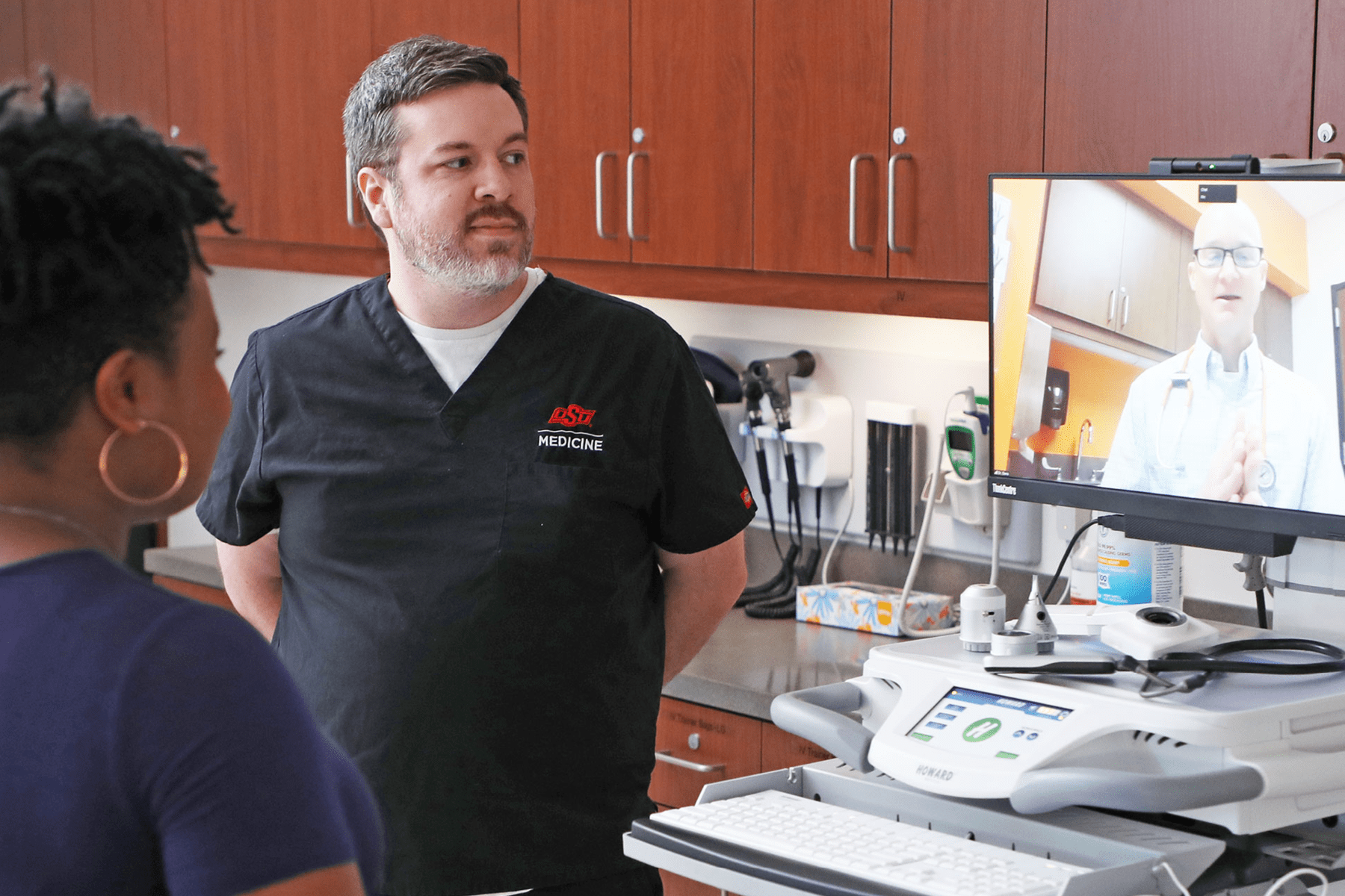 A doctor appears virtually to speak with a nurse and a patient in an examination room.