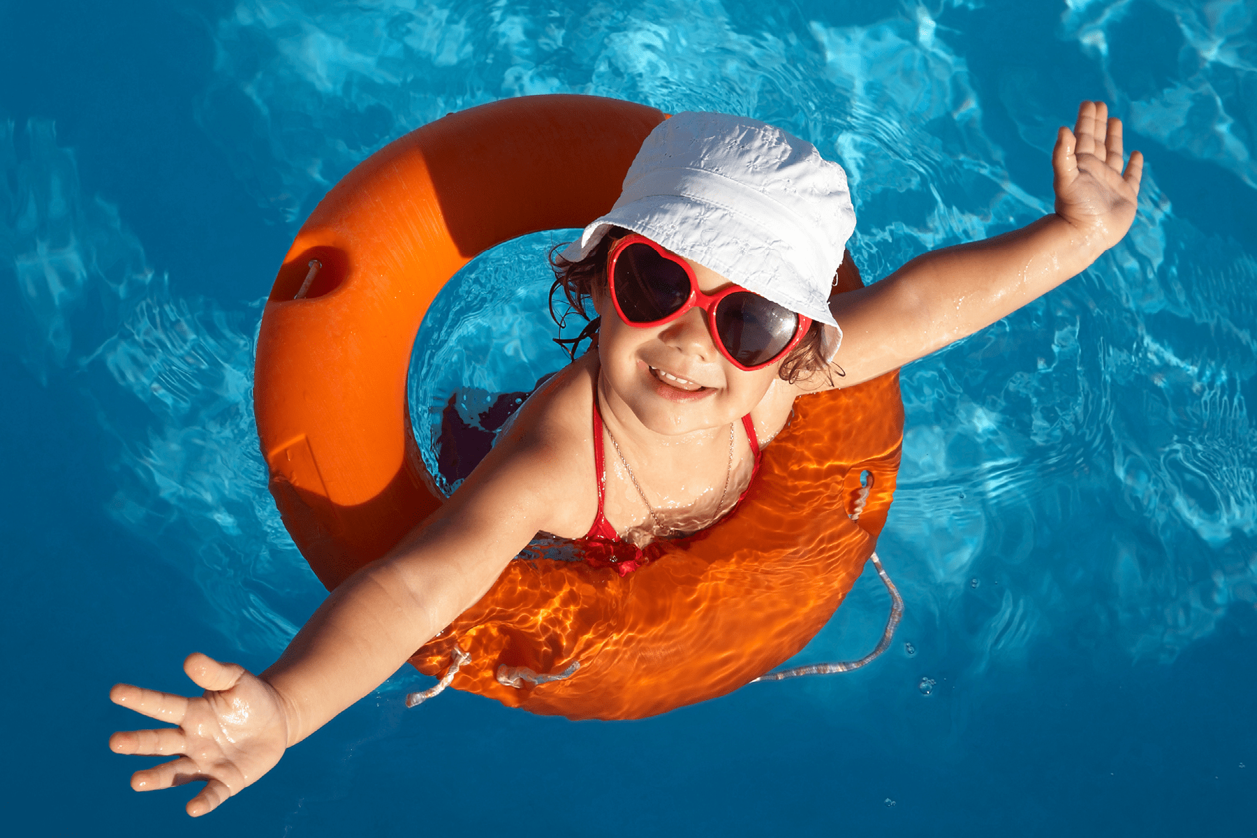 A child wearing heart shaped sun glasses wades in a pool using an orange floatation device.