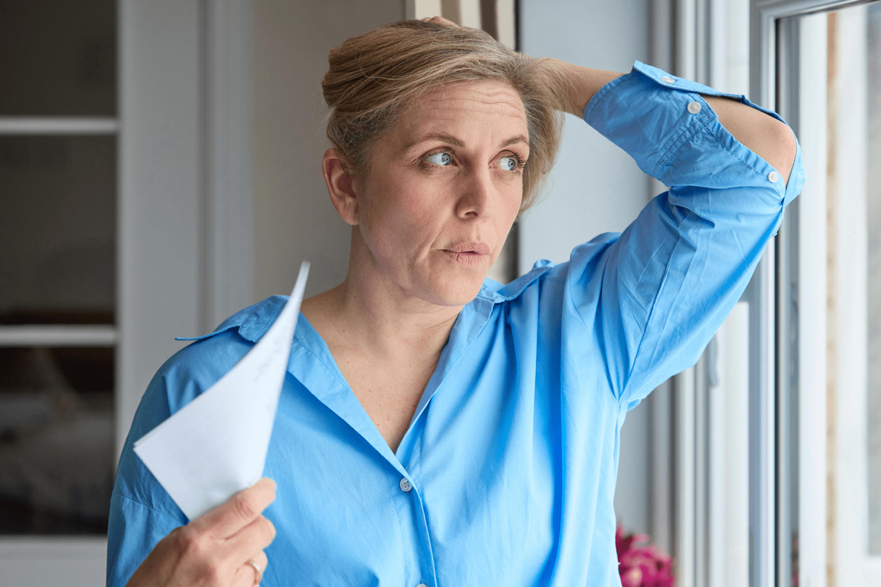 A woman looks out the window holding her hair up and fanning herself with a piece of paper.