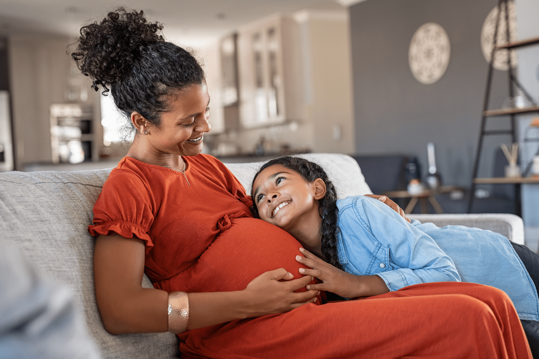 A young girl and her mother smile at each other touching the mother's pregnant belly.
