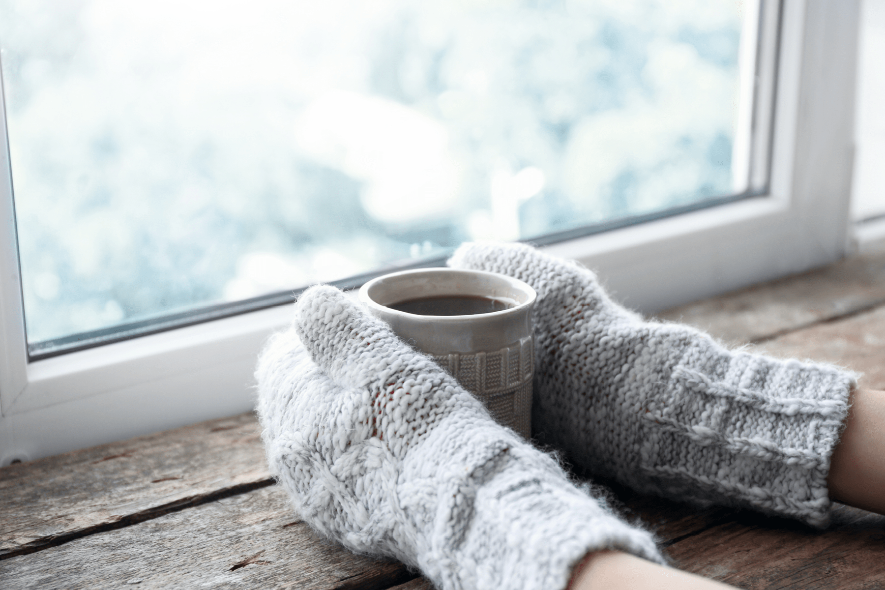 A pair of hands wearing mittens hold a cup of coffee in front of a window.