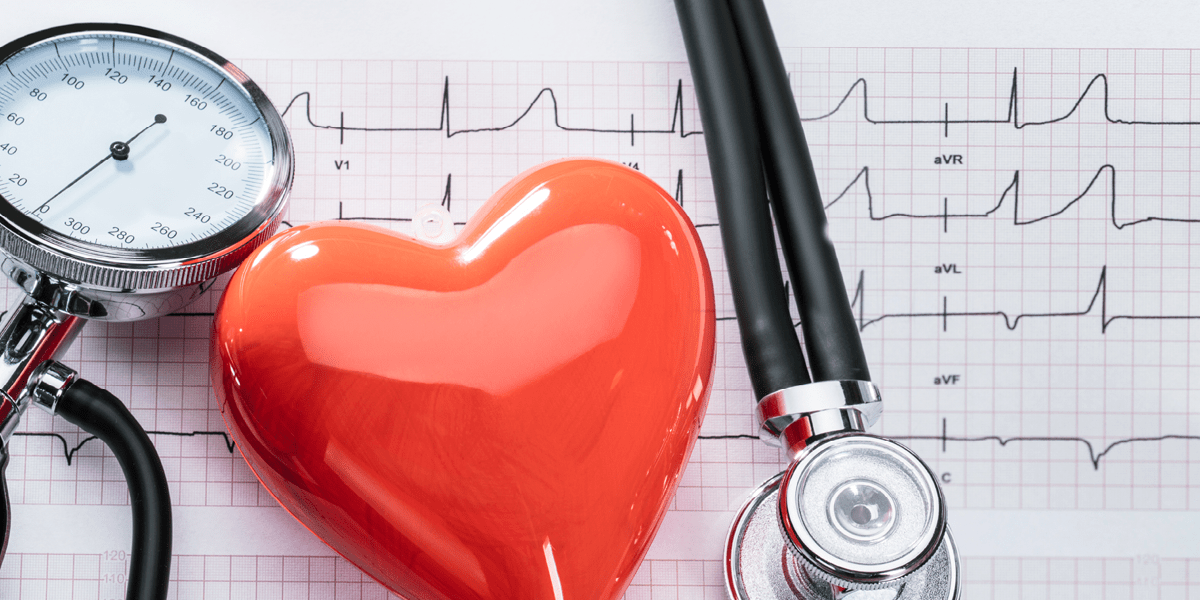 A stethoscope. the dial of a blood pressure sensor, and a shiny red heart charm sit atop a piece of graph paper with heart rate monitoring lines drawn of it.