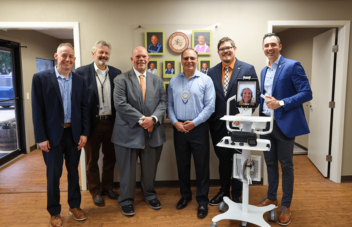 antlers-clinic-news Six people appear in person, and one digitally via a tablet device attached to a portable cart in a group before the Great Seal of the Choctaw Nation Smiling.
