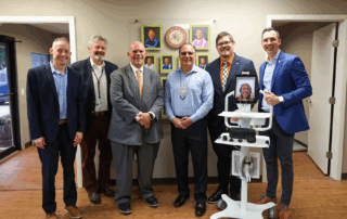 Six people appear in person, and one digitally via a tablet attached to a portable cart in a group before the Great Seal of the Choctaw Nation Smiling.