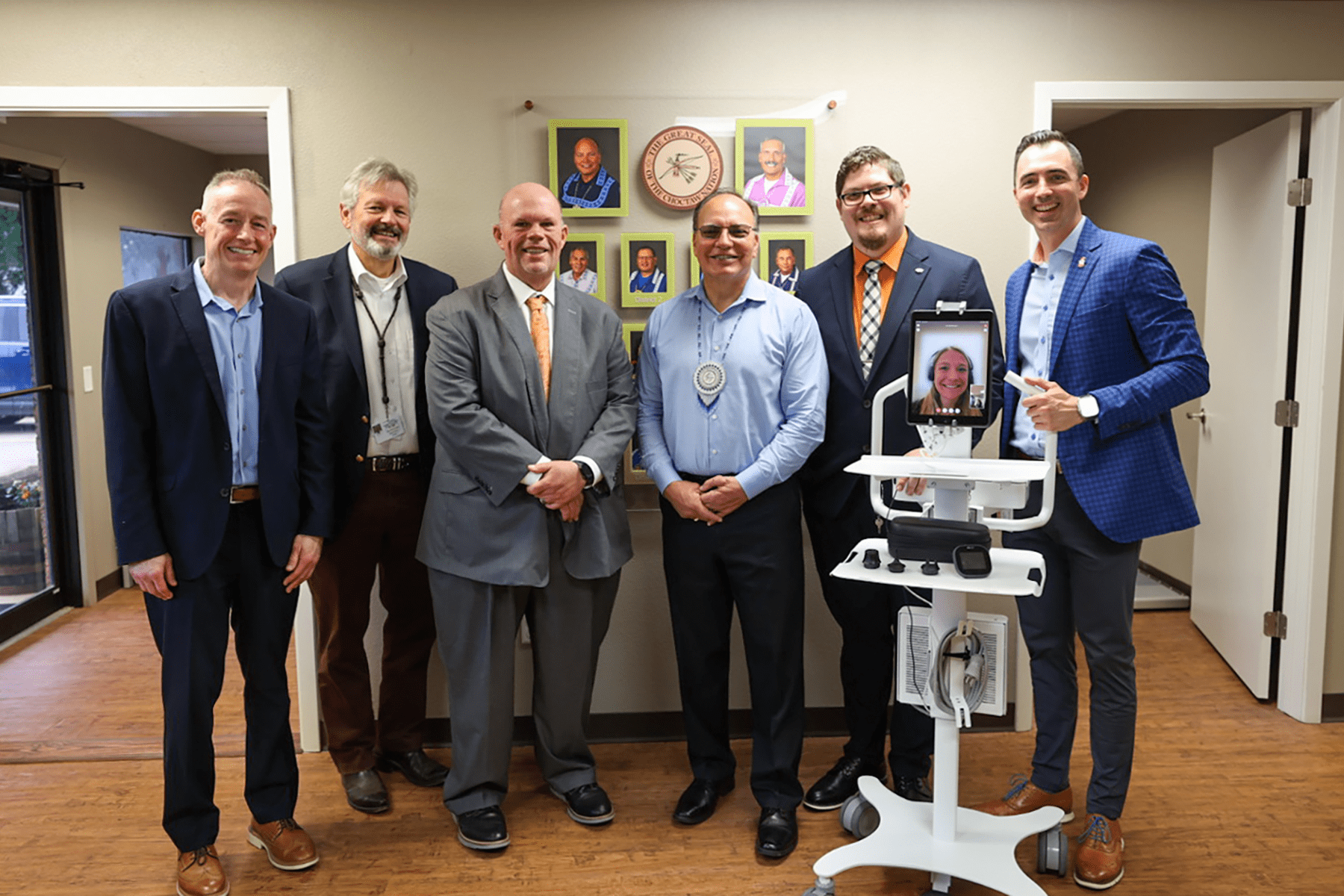 Six people appear in person, and one digitally via a tablet attached to a portable cart in a group before the Great Seal of the Choctaw Nation Smiling.